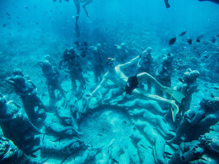 Man underwater sitting at bottom of sea with statues around him