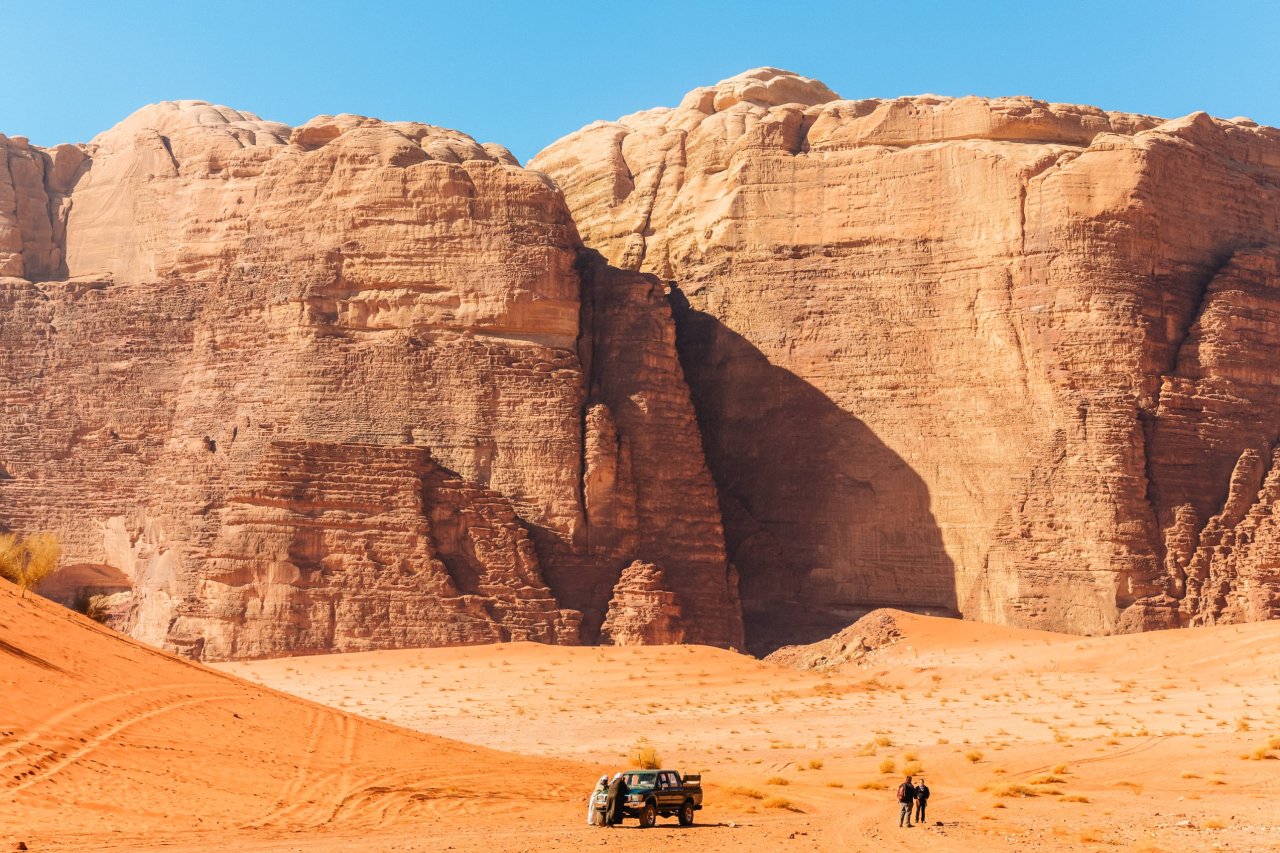 Wadi Rum desert with a huge rock face in the distance. In the front of the rock is a jeep with tow young travellers taking a photo.