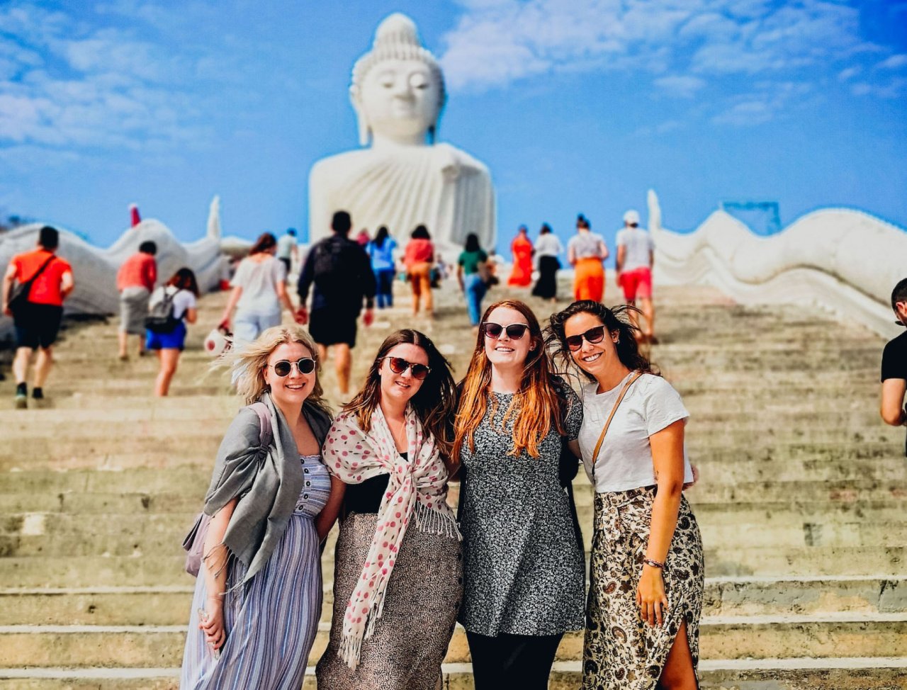 Group of girls standing at the bottom of steps with big white Buddha behind them and crowds of people visiting the site.